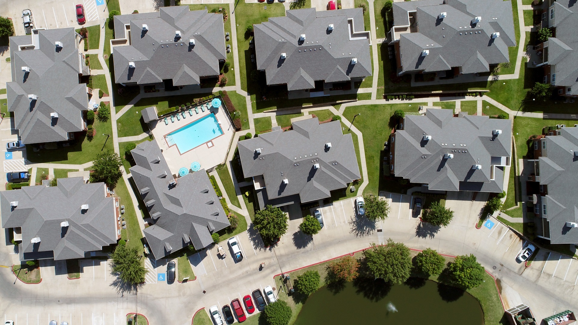 Arial shot of apartments where only the roofs are visible with paths between buildings, green grass, trees, and the community area with the pool